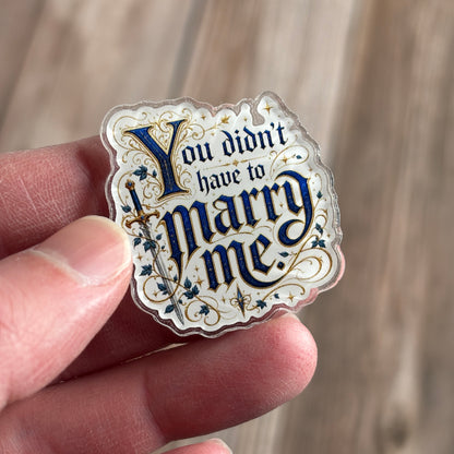 Die-cut holographic acrylic pin held between fingers, illuminated-manuscript-style navy and gold text "You didn't have to marry me." beside an ivy-wrapped sword.